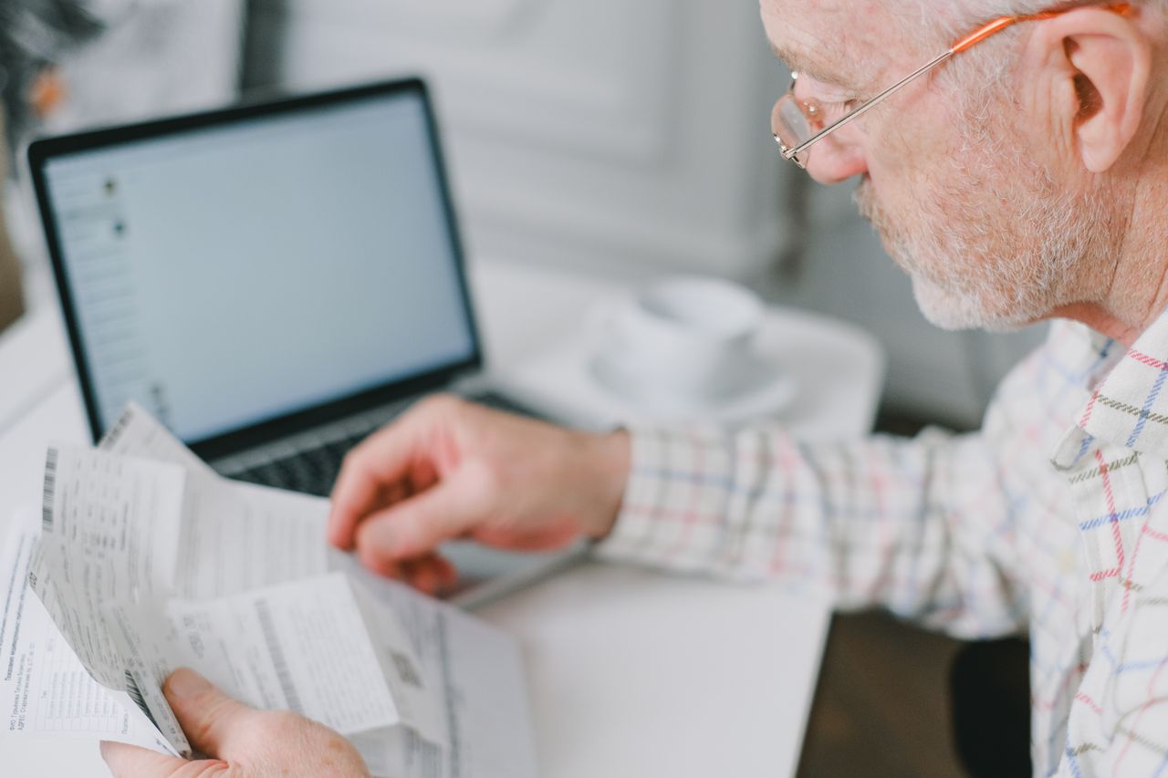 gallery-3 Elderly man wearing eyeglasses reading documents next to a laptop at home.