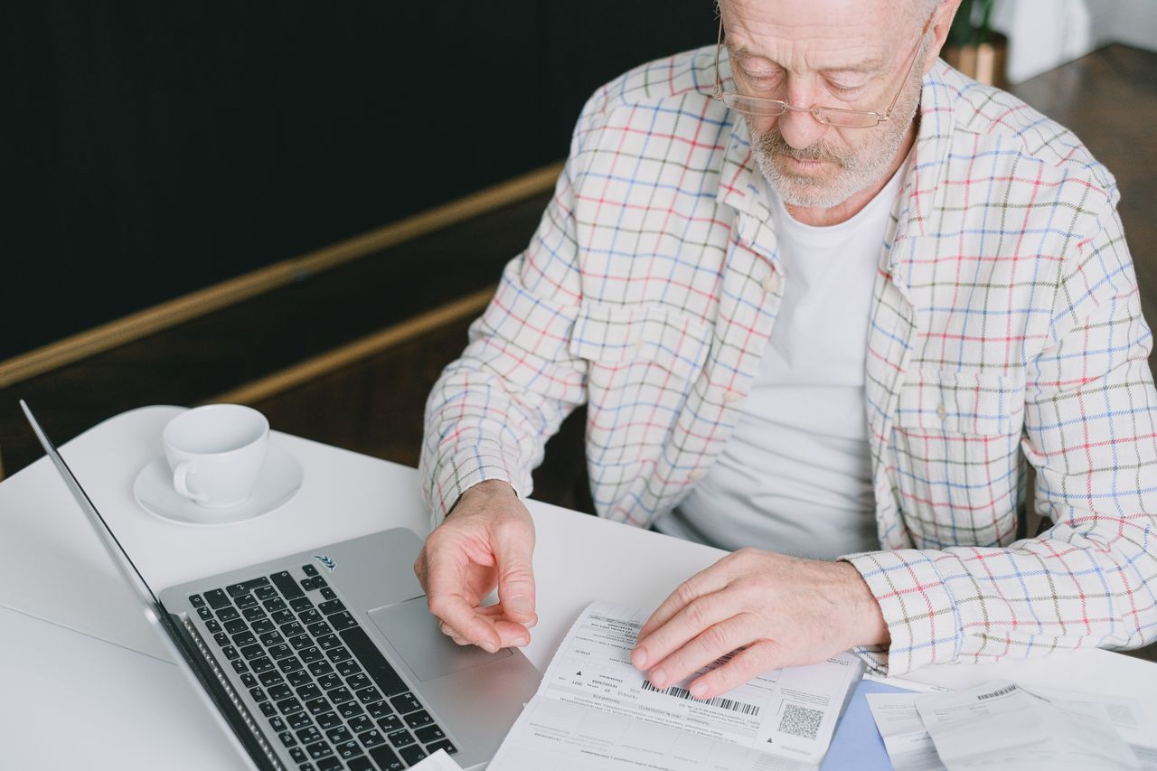 gallery-4 Elderly man with eyeglasses reviewing documents at a laptop. Indoor setting with natural light.